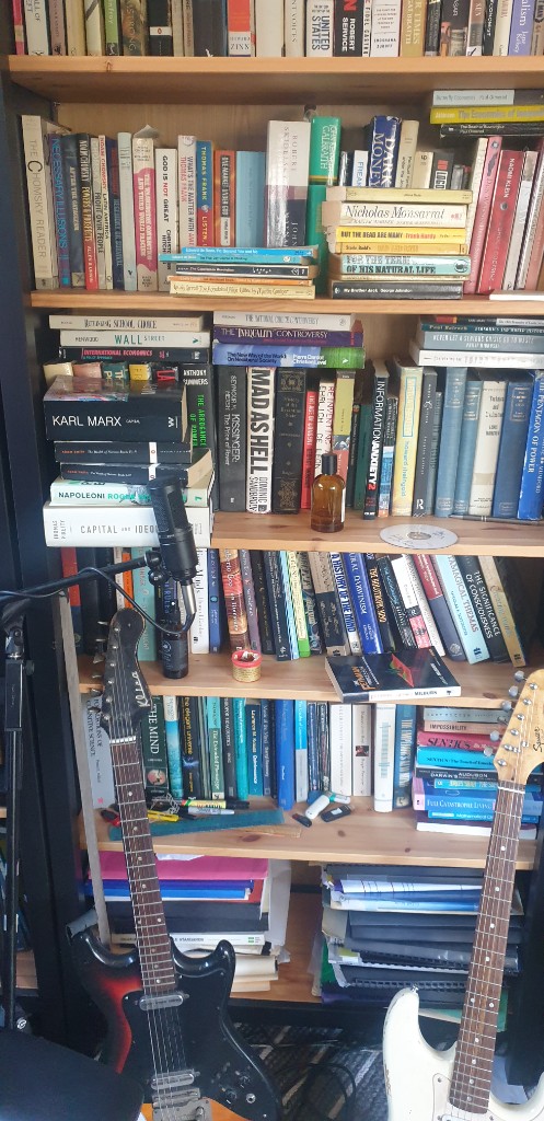 Floor-to-ceiling bookshelf with political and economics non-fiction, two electric guitars, and a studio microphone in the foreground.